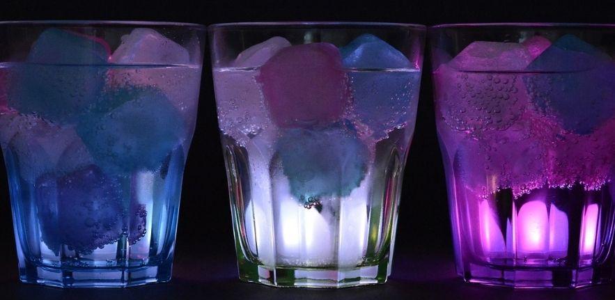 three glasses of water containing ice cubes and pink and blue lights going through them from the back. 
