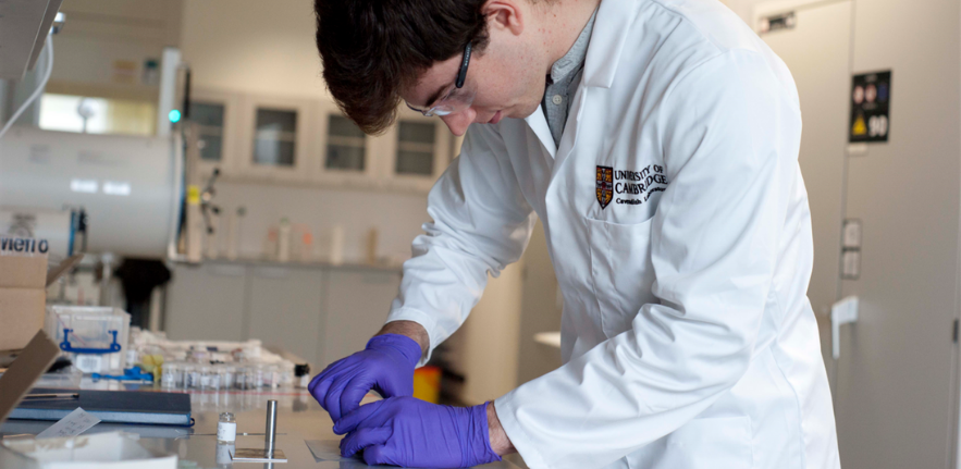 Young man wearing a lab coat, protective goggles and purple gloves working at a lab bench. 