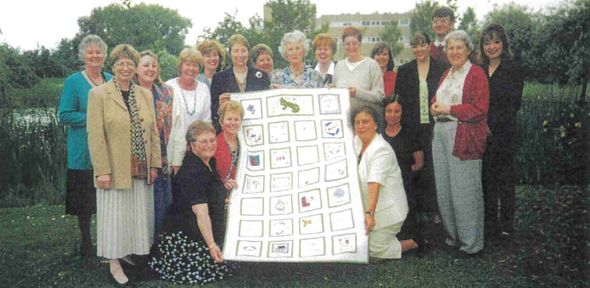 A large group of women standing oustide and showing a handmade throw. Catherine Clifton is kneeling and holding the thow on the right side. She's wearing a white suit.